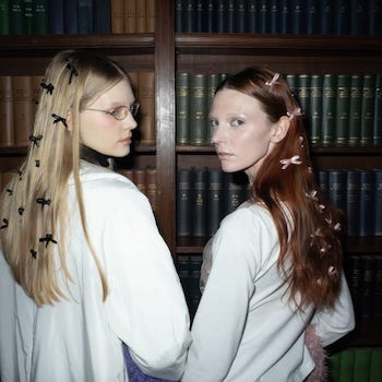 two girls wearing several bows in their hair in front of a bookshelf with their bodies turned away, facing the camera 